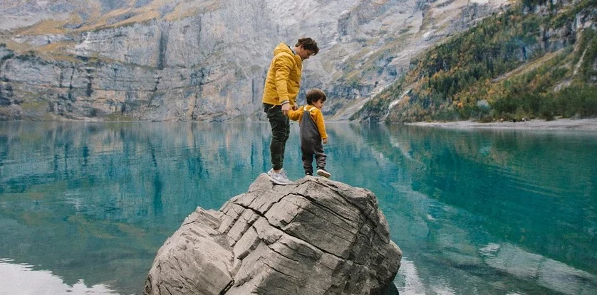 A parent and child standing on a rock above a turquoise mountain lake
